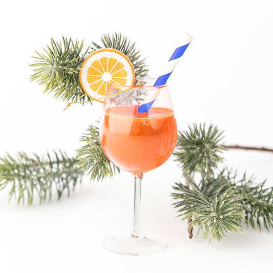 Aperitif in a stemmed glass with a straw and orange slice, surrounded by pine branches on a white background