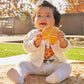Child holding a yellow teether toy outdoors with trees in the background