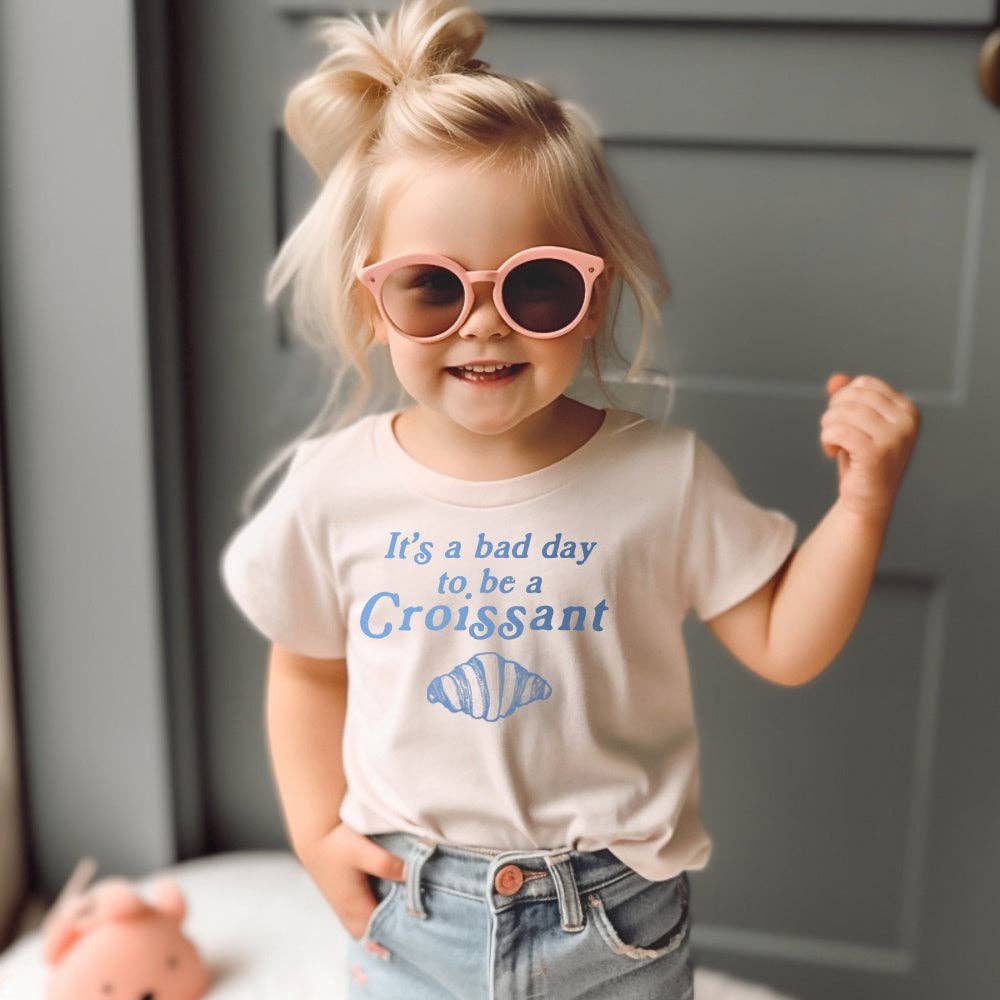 Child wearing pink sunglasses and a t-shirt with a humorous message "it's a bad day to be a croissant" 