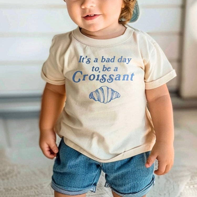 Child wearing a t-shirt with text and a  croissant graphic, standing indoors.