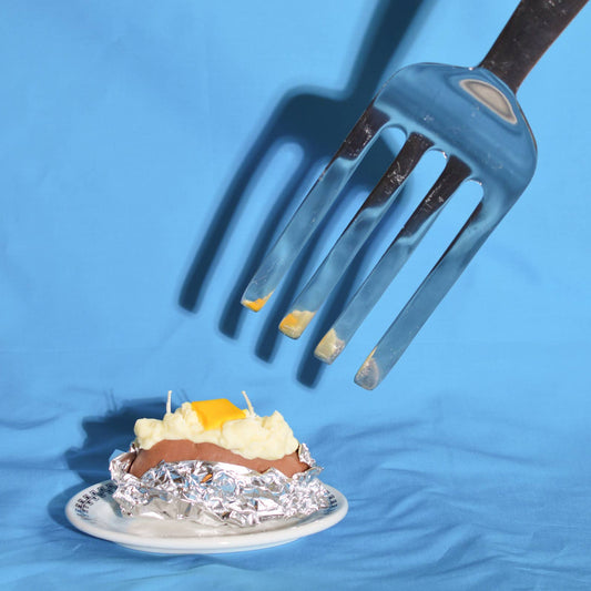 candle that looks like a baked potato with a pad of butter on top and a giant fork coming from the top right corner, about to cut into it