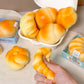 Stress balls that look like baked goods in a white container with a hand holding a croissant stress ball on a light surface.