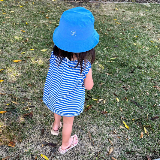 Child wearing a blue striped dress and blue bucket hat standing on grass with scattered leaves.