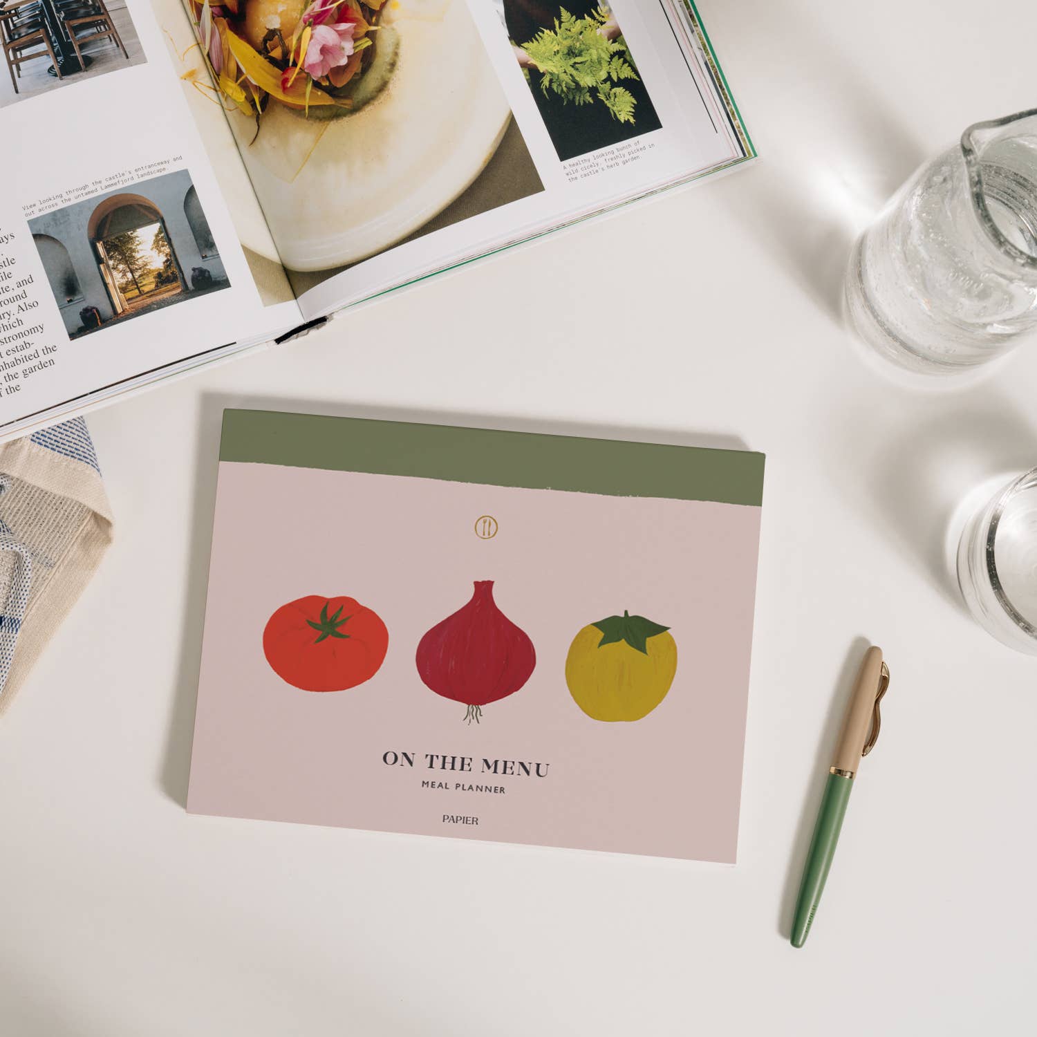Front cover of Vegetable Medley Meal Planner. Features illustration of tomato, red onion and yellow apple on neutral background with rustic green stripe along the spine. Cover text reads "ON THE MENU... meal planner. " Shown on white table with magazine and pen.