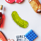 A pickle magnet photographed with a bottle of campari and colorful objects on a white background