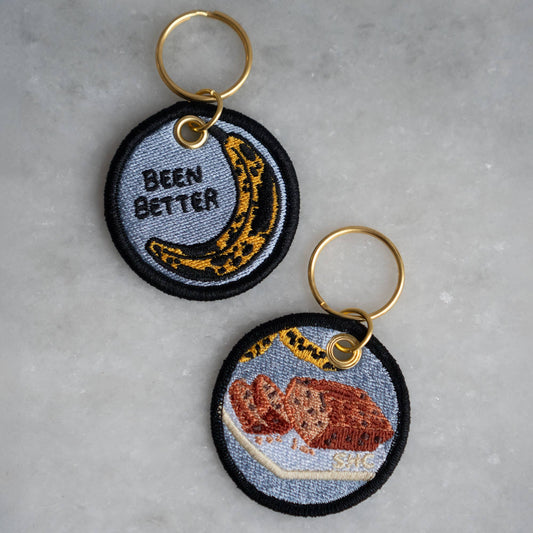 two sides of a keychain, each embroidered with a different design. The top one has a bruised banana on it with black text that reads "Been Better" while the other has banana bread embroidered on it