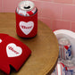 Red 'Beer' can coolers with a heart design on a wooden table against a pink tiled wall.