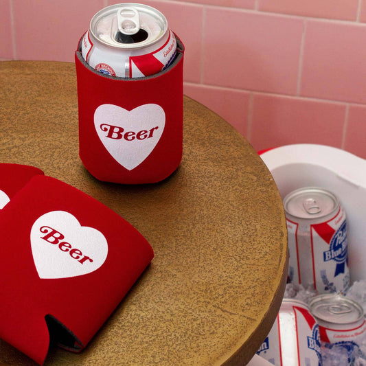 Red 'Beer' can coolers with a heart design on a wooden table against a pink tiled wall.