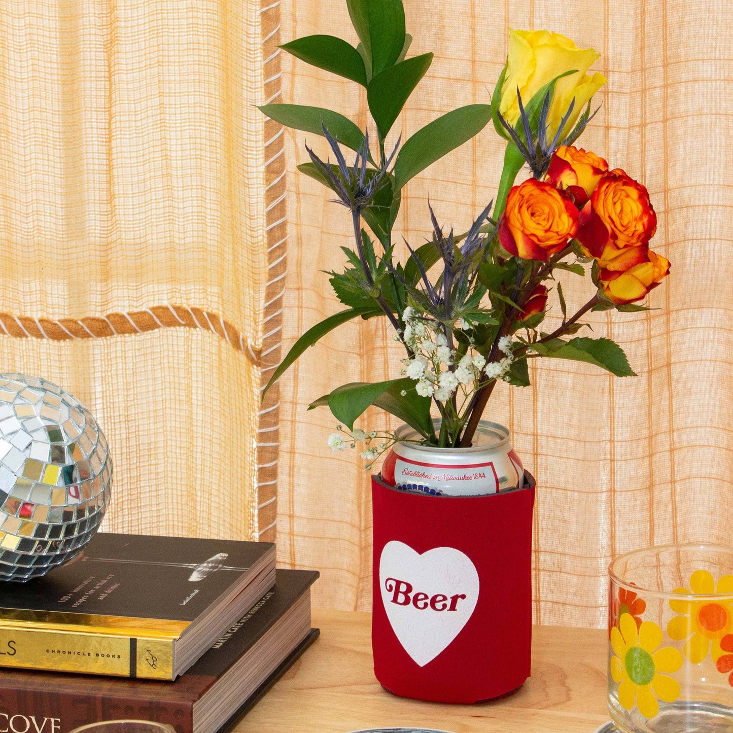 Red can cooler with 'Beer' heart design on a table with books, flowers, and glasses.