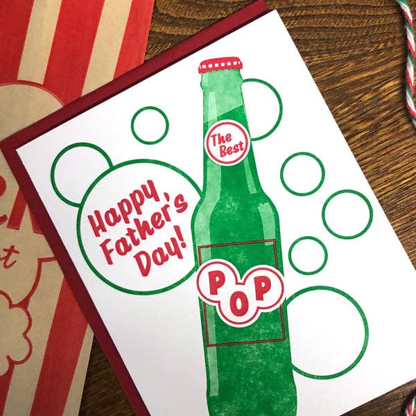 Father's Day card with green bottle design and 'Happy Father's Day!' text on a wooden surface.