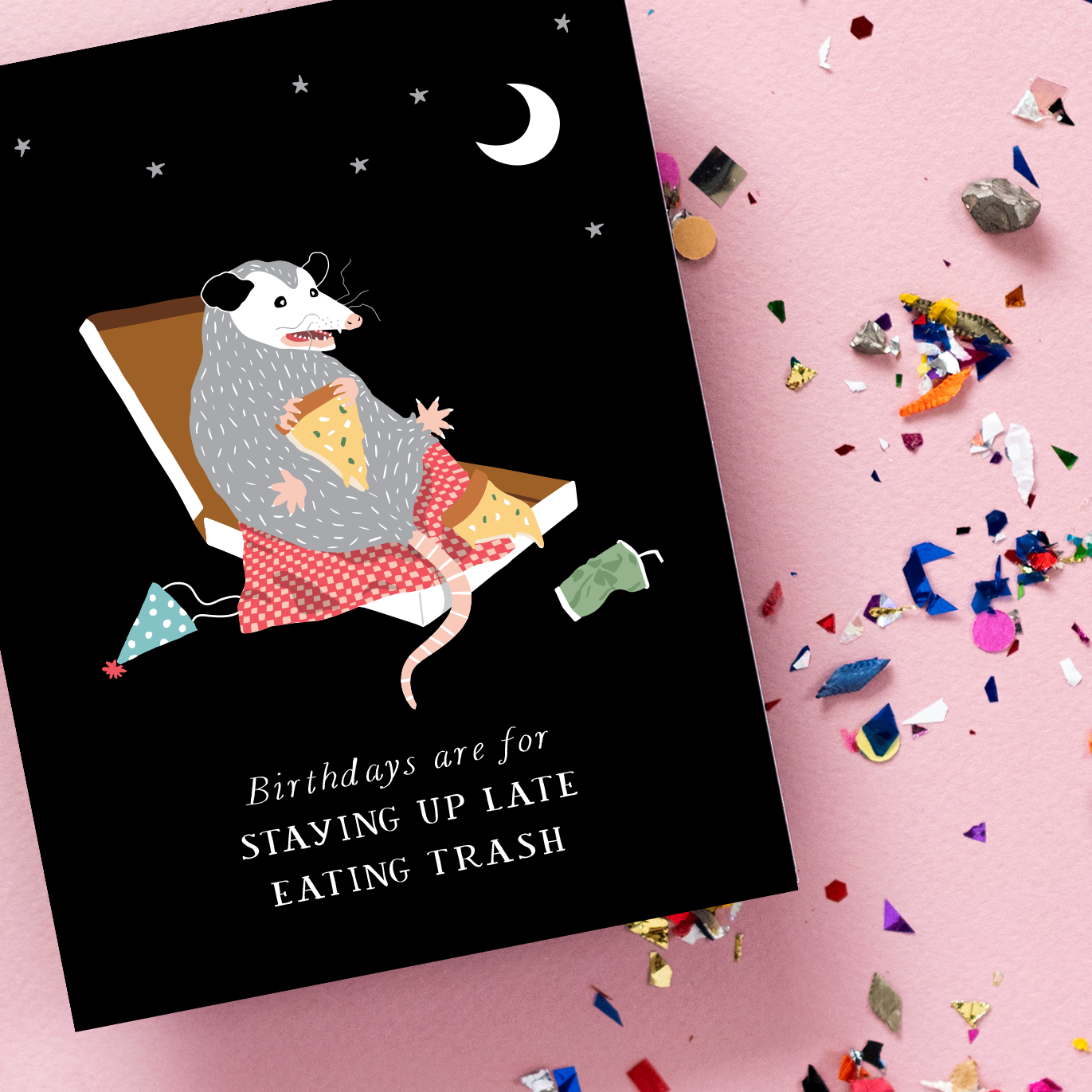 Card with an opposum illustration and text 'Birthdays are for staying up late eating trash' on a black background, with confetti on a pink surface.