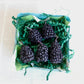 Decorative box filled with green paper shreds and blackberry shaped candles on a light background