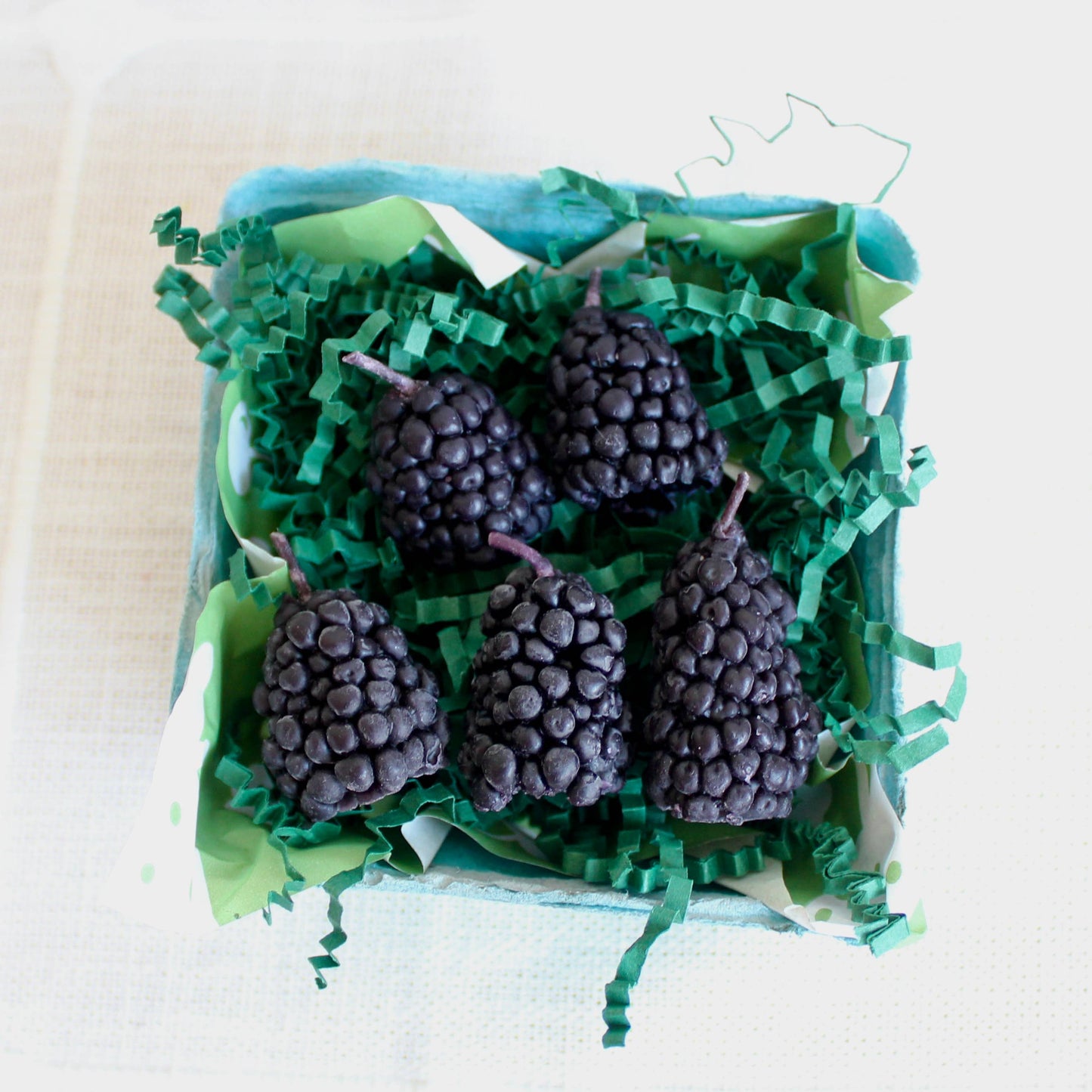 Decorative box filled with green paper shreds and blackberry shaped candles on a light background