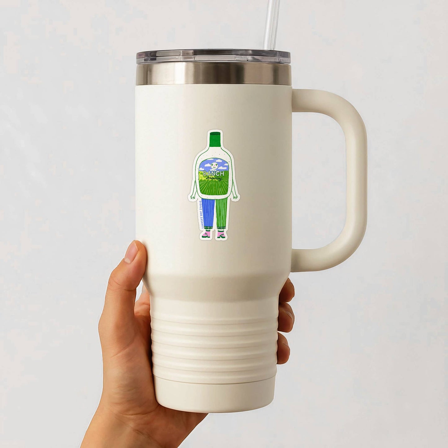 White insulated mug with a handle held by a hand, featuring a colorful sticker on a light gray background