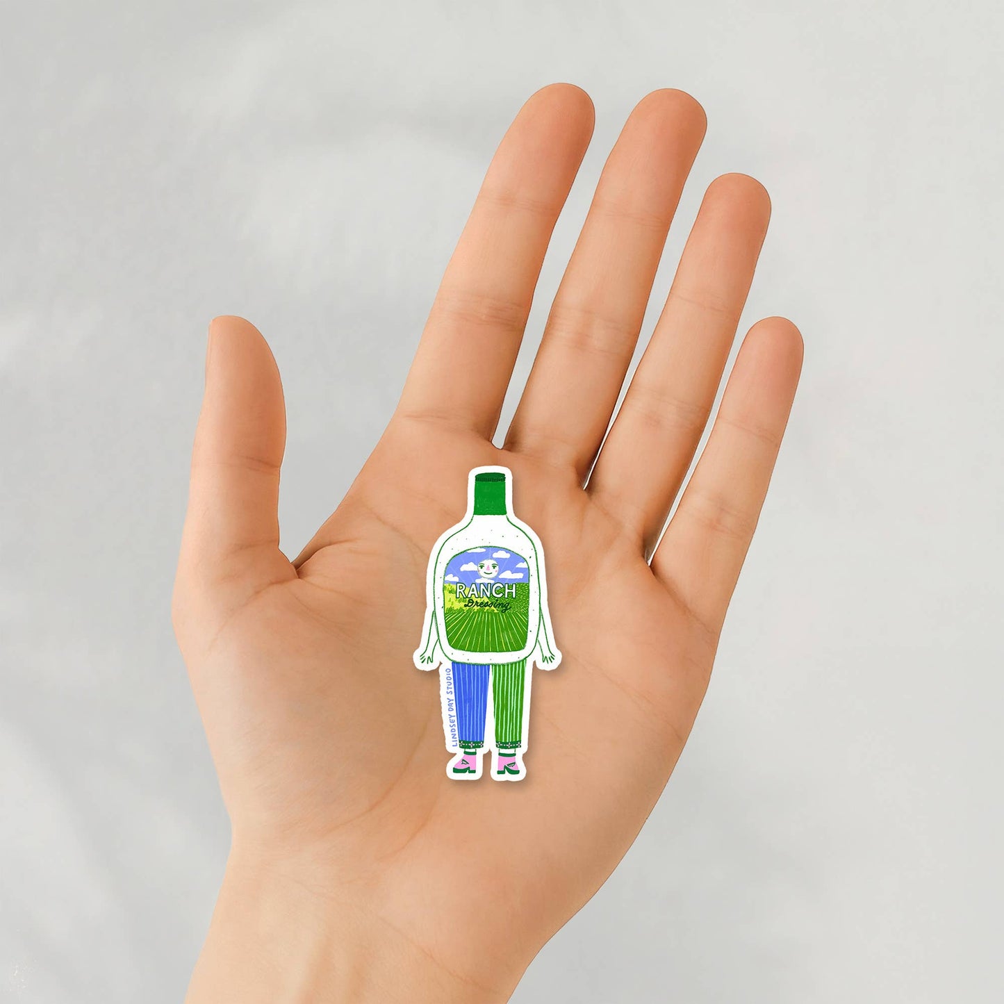 Hand holding a bottle-shaped sticker with a green label against a light gray background