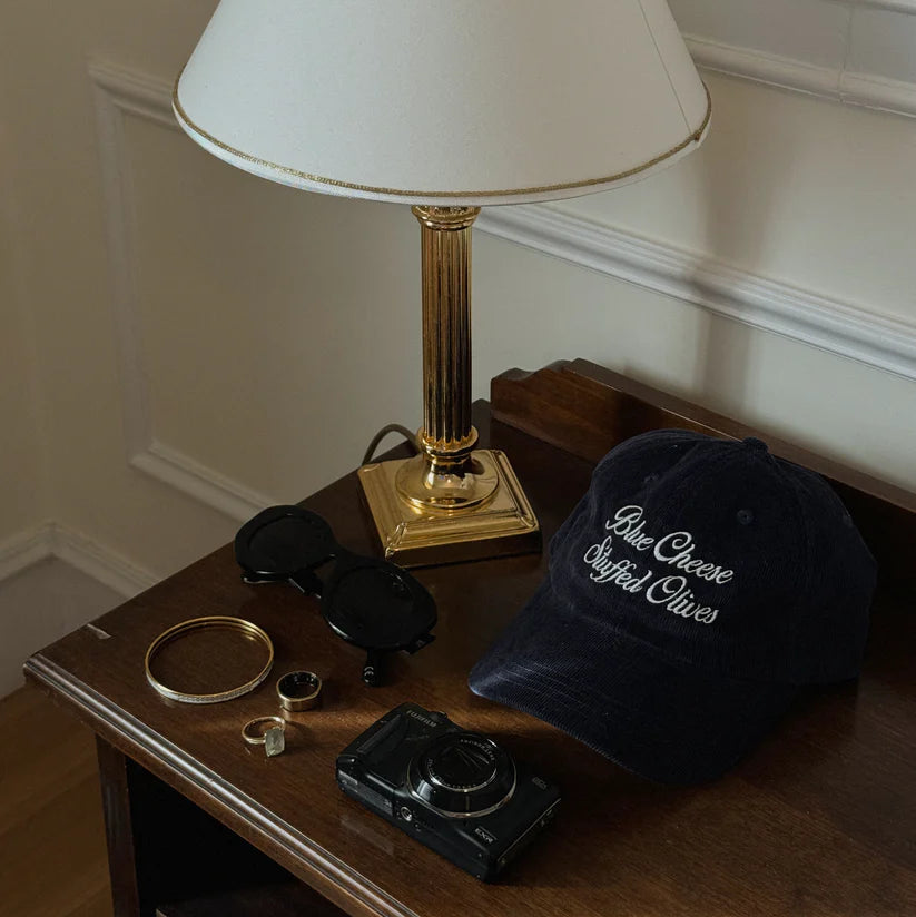 Blue corduroy hat with "blue cheese stuffed olives" text on a table with keys, jewelry, sunglasses and a lamp