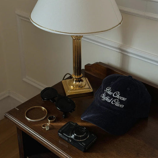 Blue corduroy hat with "blue cheese stuffed olives" text on a table with keys, jewelry, sunglasses and a lamp