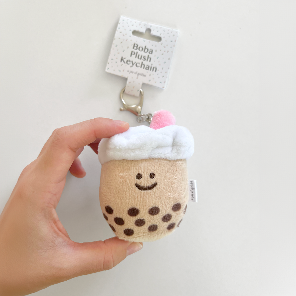 Plush keychain shaped like a bubble tea with a smiling face, held by a hand against a plain background.