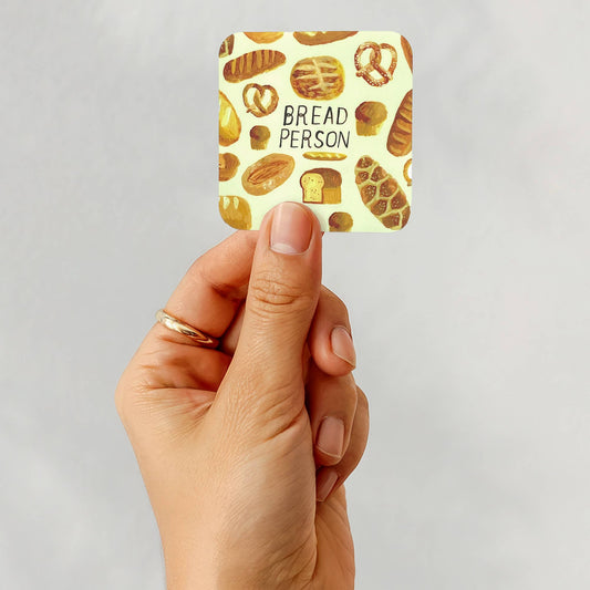 A hand holding a square sticker featuring a pattern of various breads and the text 'BREAD PERSON' in the center, set against a plain light gray background.