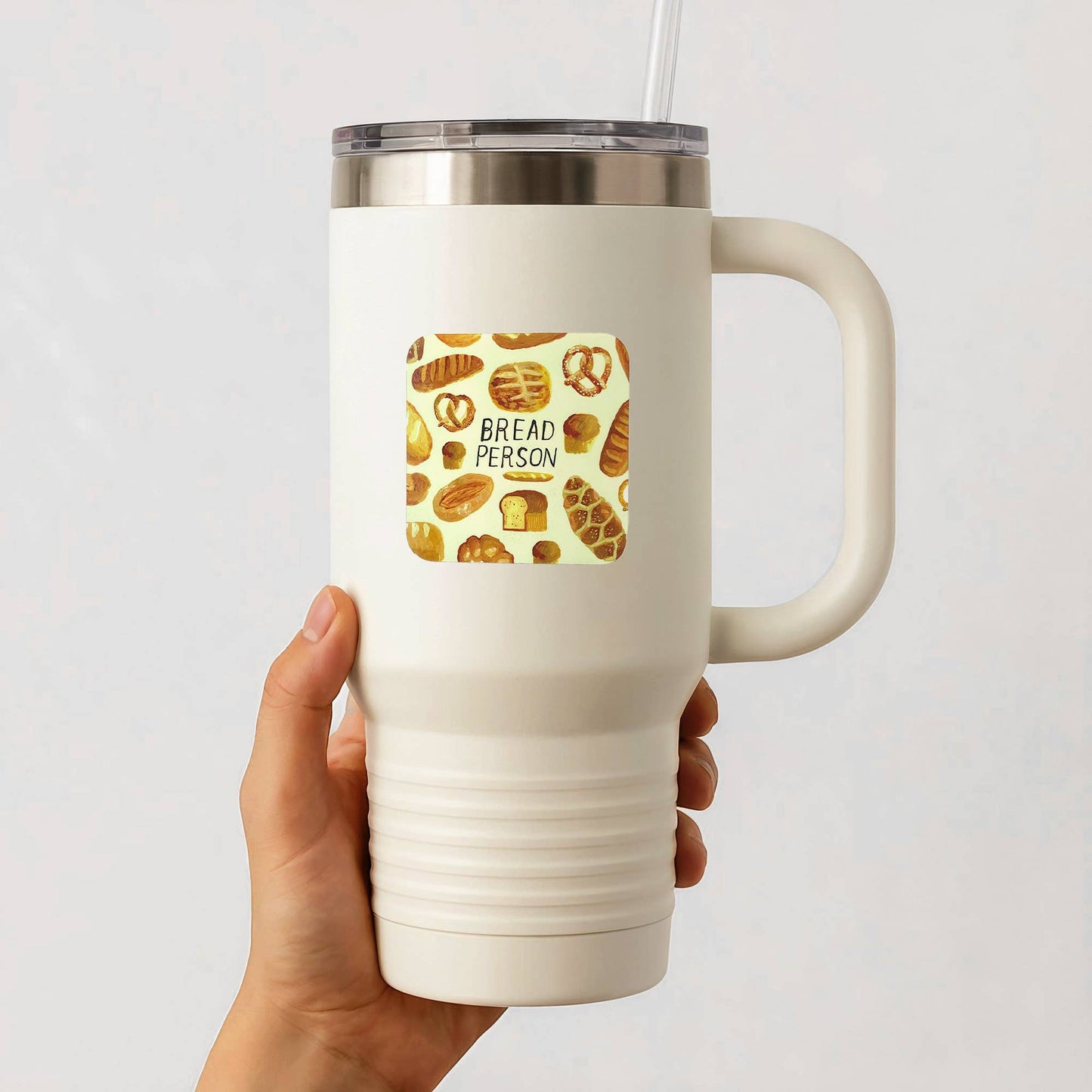 A hand holding a white insulated travel tumbler with a handle, clear lid, and straw. The tumbler has a square graphic on the front featuring illustrations of various breads and the text 'BREAD PERSON'.