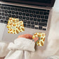 A hand wearing a light gray sweatshirt holds a small square sticker with a pretzel and cinnamon bun pattern next to two matching stickers applied to a laptop trackpad. The scene is styled on a white faux-fur surface with a laptop and disco balls in the background.