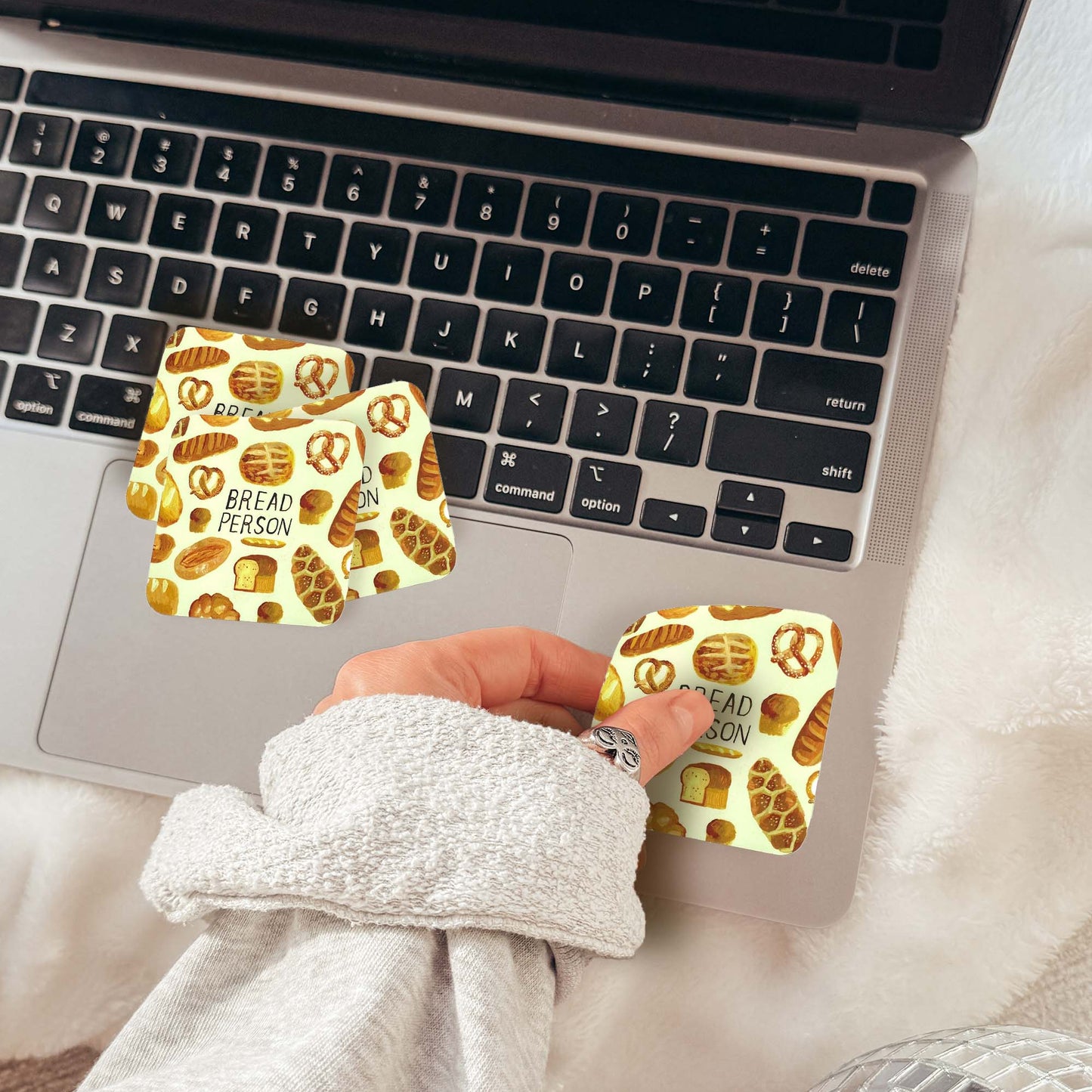 A hand wearing a light gray sweatshirt holds a small square sticker with a pretzel and cinnamon bun pattern next to two matching stickers applied to a laptop trackpad. The scene is styled on a white faux-fur surface with a laptop and disco balls in the background.