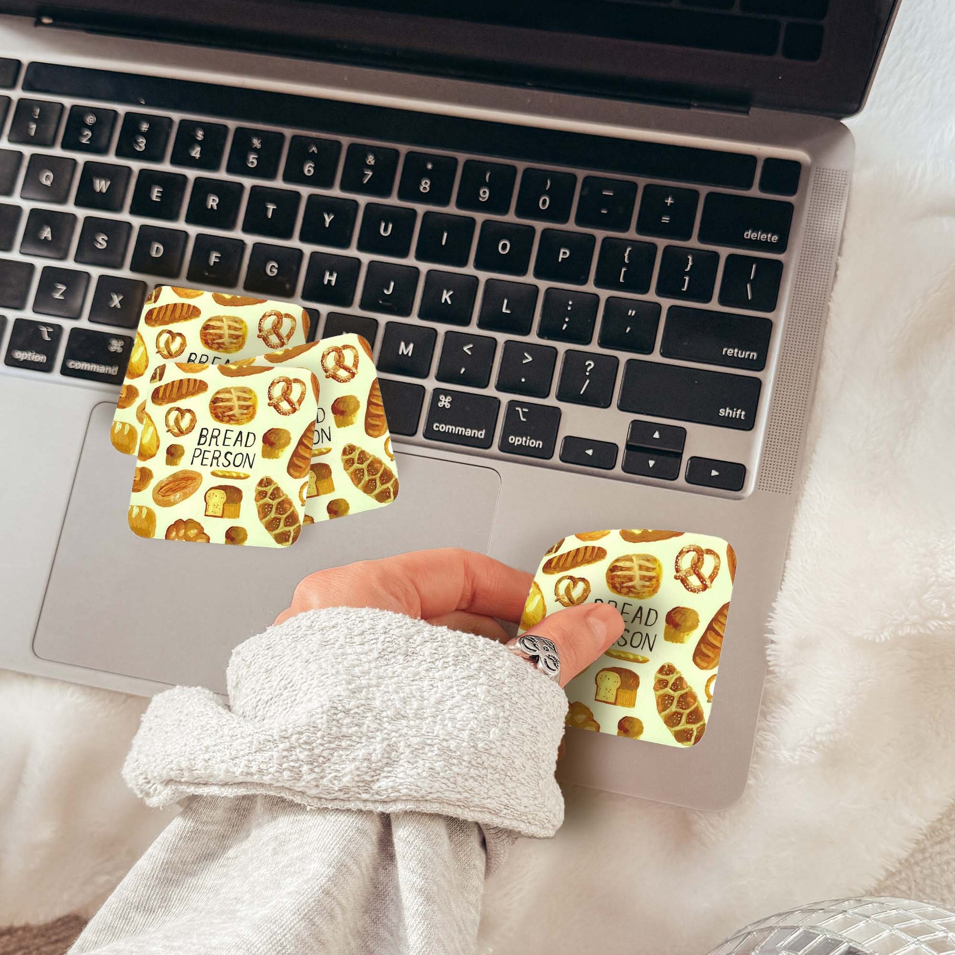 A hand wearing a light gray sweatshirt holds a small square sticker with a pretzel and cinnamon bun pattern next to two matching stickers applied to a laptop trackpad. The scene is styled on a white faux-fur surface with a laptop and disco balls in the background.