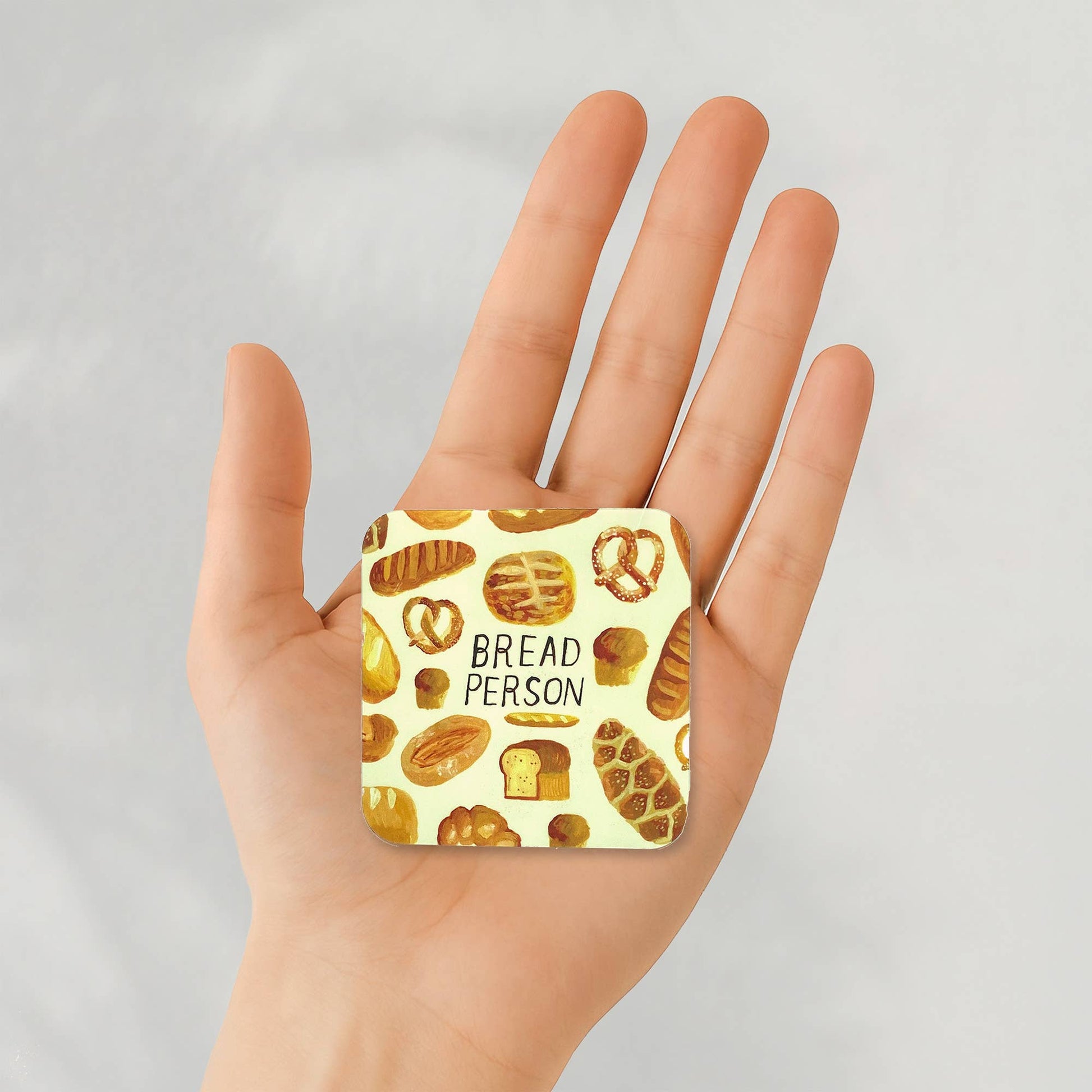A hand holding a square sticker with rounded corners featuring a light yellow background and a pattern of various breads, loaves, and pretzels, with the text 'BREAD PERSON' printed in black.