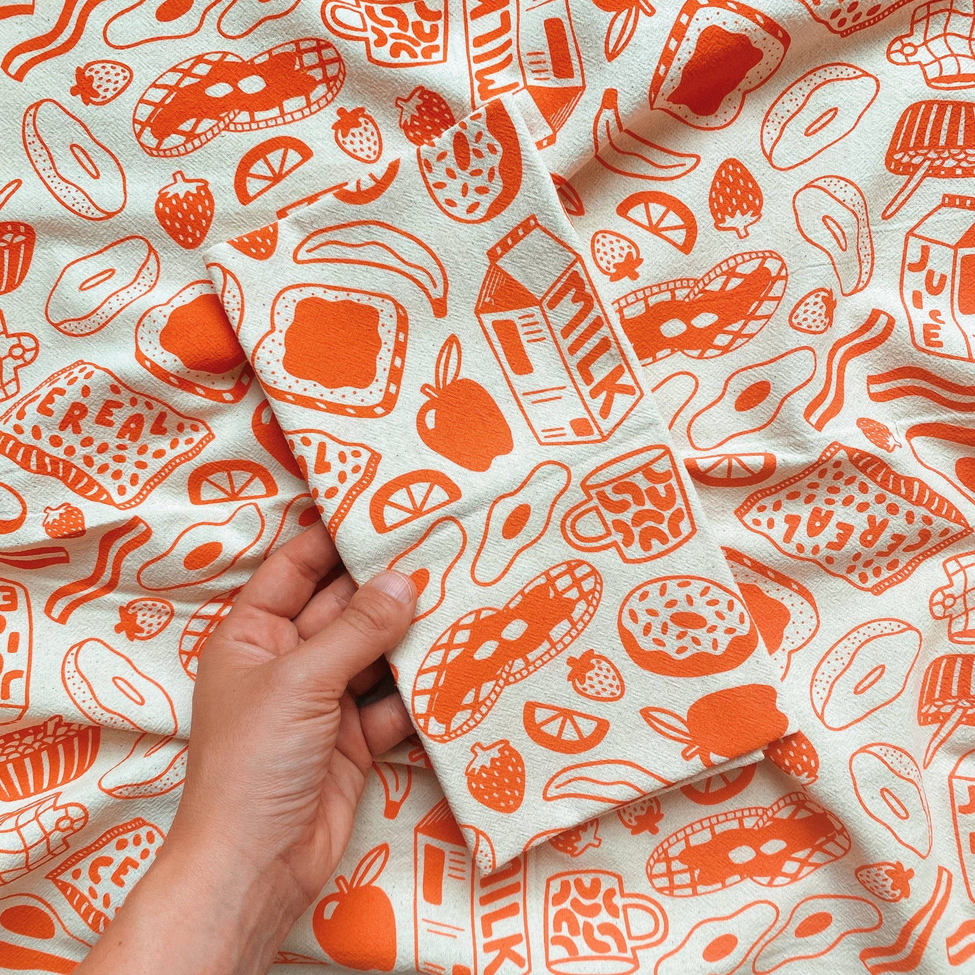 Kitchen towel with various breakfast food and drink items such as toast, fruits, eggs, waffles, donuts milk and a mug. Printed in orange ink on natural colored fabric