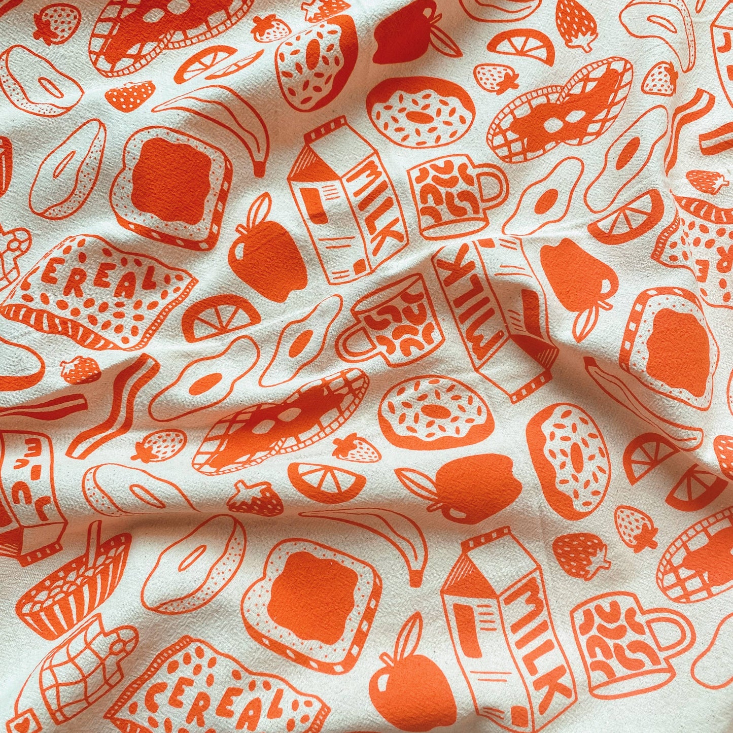 kitchen towel with various breakfast food and drink items such as toast, fruits, eggs, waffles, donuts milk and a mug. Printed in orange ink on natural colored fabric