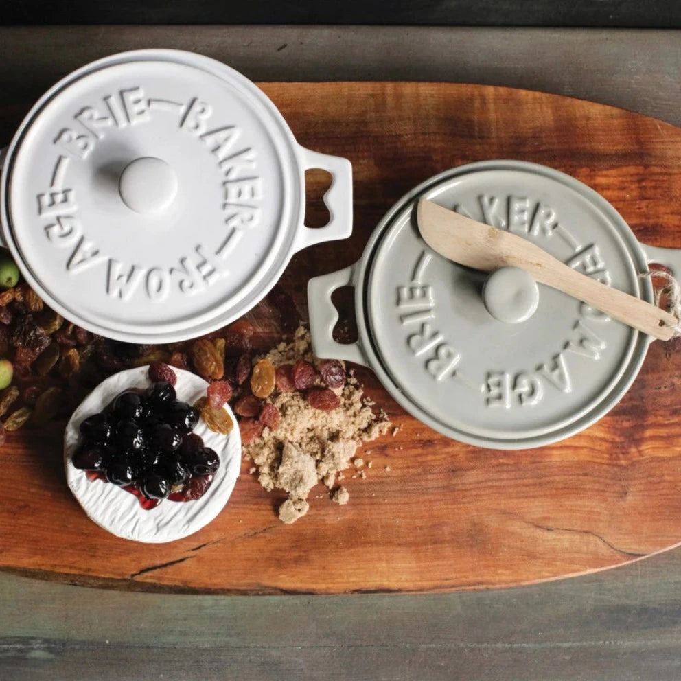 Wood platter with 1 white and 1 gray brie baker. The board has staged dried fruit, brown sugar, and a brie with cherries on top. Top view.