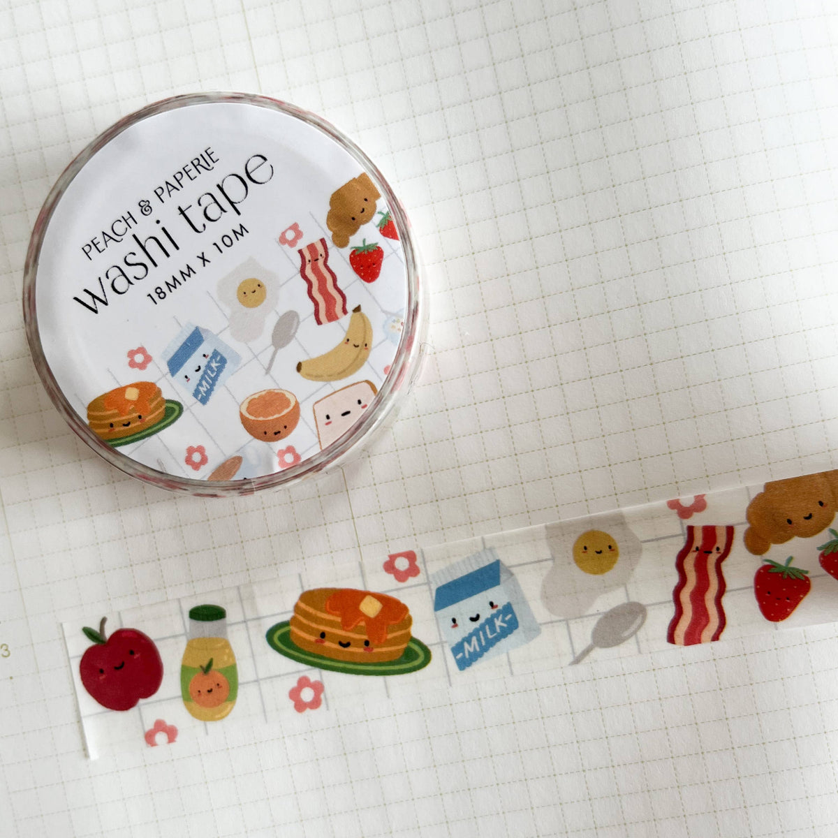 A roll of Peach & Papirie washi tape with a cute breakfast-themed pattern, shown next to a strip of the tape unrolled on a white grid notebook.