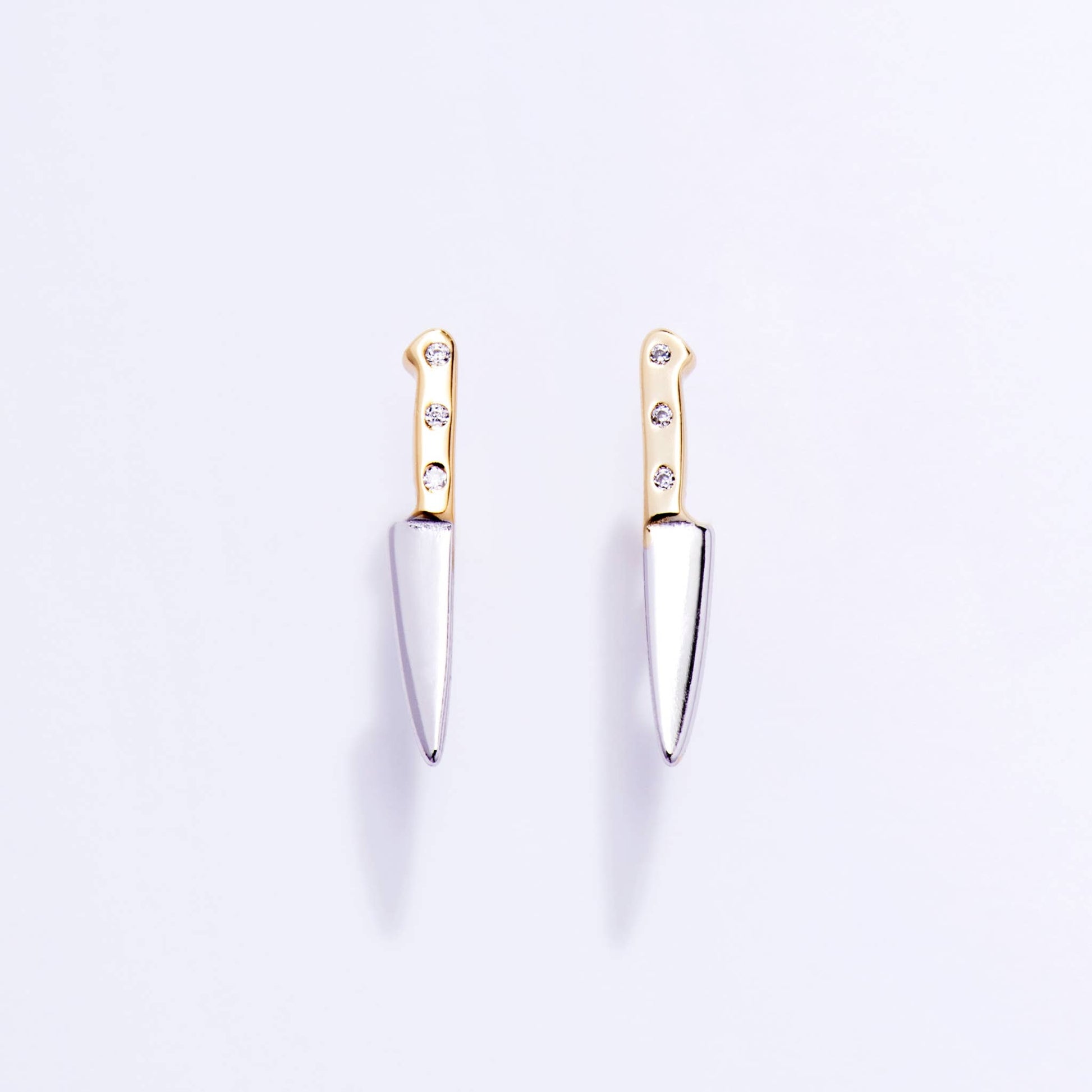 Pair of knife-shaped earrings with gold handles and silver blades on a light purple background