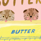 Illustration of two cats peeking over a stick of butter against a pink background.