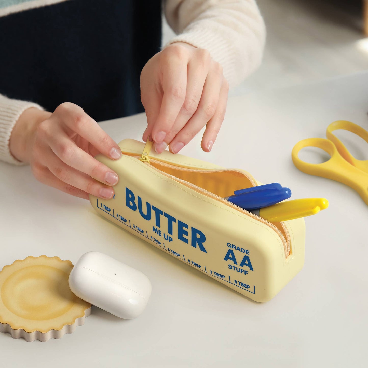 Person opening a yellow pencil case with 'BUTTER' branding on a light surface.
