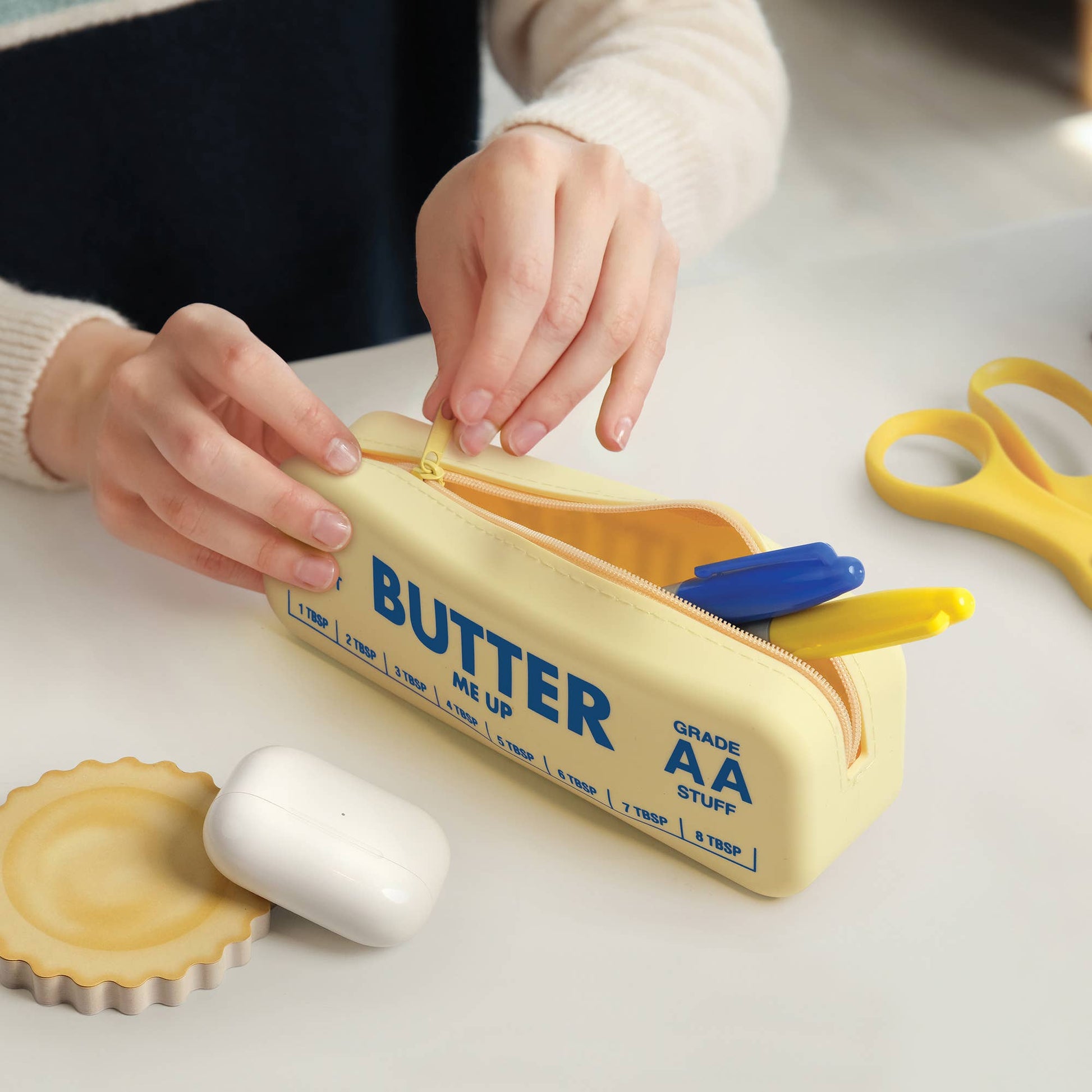 Person opening a yellow pencil case with 'BUTTER' branding on a light surface.