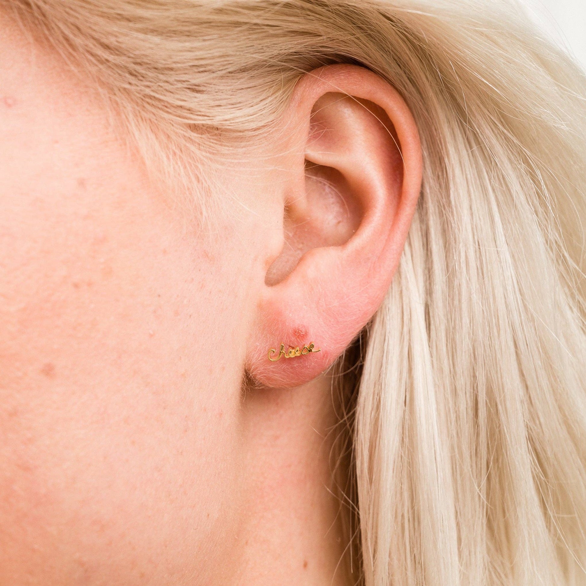 Single 14k gold plated stud earring -- reads "cheese" in handwritten script. Worn on ear for scale 