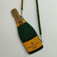 Photo of beaded purse shaped like a champagne bottle hanging from a green and gold beaded strap. Displayed on a plain white background. Beaded design depicts green bottle with yellow and blue nondescript wine label and gold foil at the top of bottle.