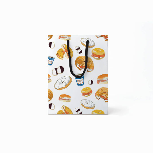 Gift bag with black rope handles and designed with various illustrations of black & white cookies, coffee in a blue paper cup, bagels with schmear or lox 