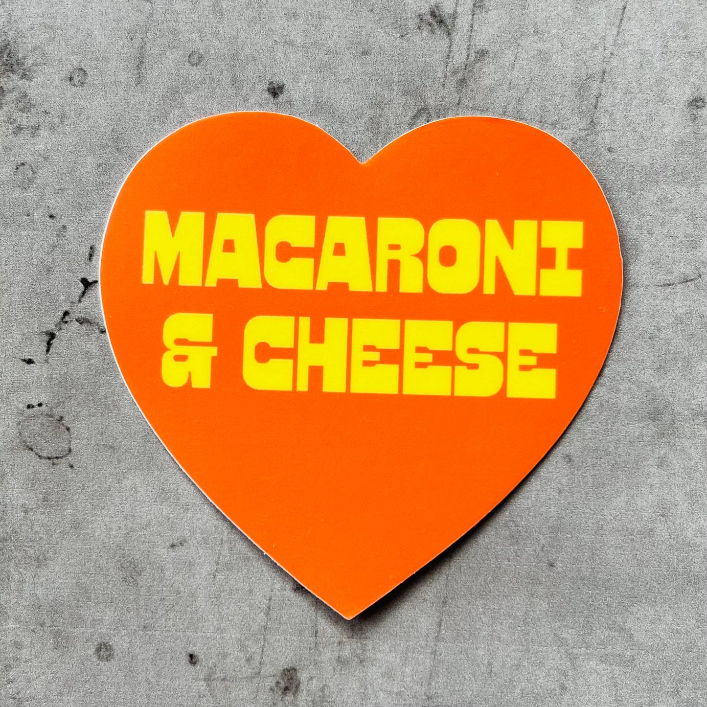 Orange, heart-shaped sticker with "macaroni & cheese" printed in quirky yellow font.