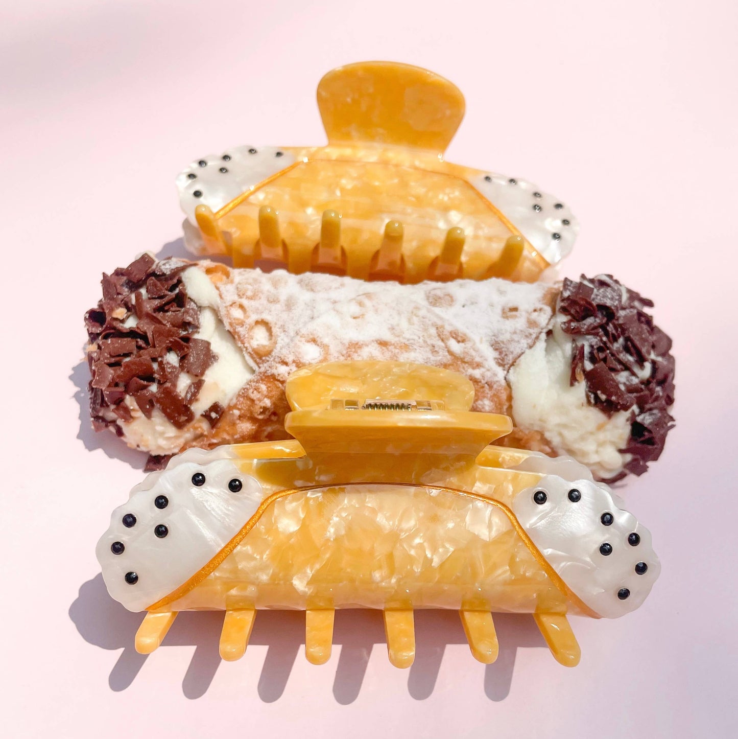 Two cannoli-shaped hair clips with polka dot details to mimic chocolate chips on a light pink background
