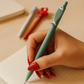 Hand holding a green fish-shaped pen over a notebook with other pens in the background.