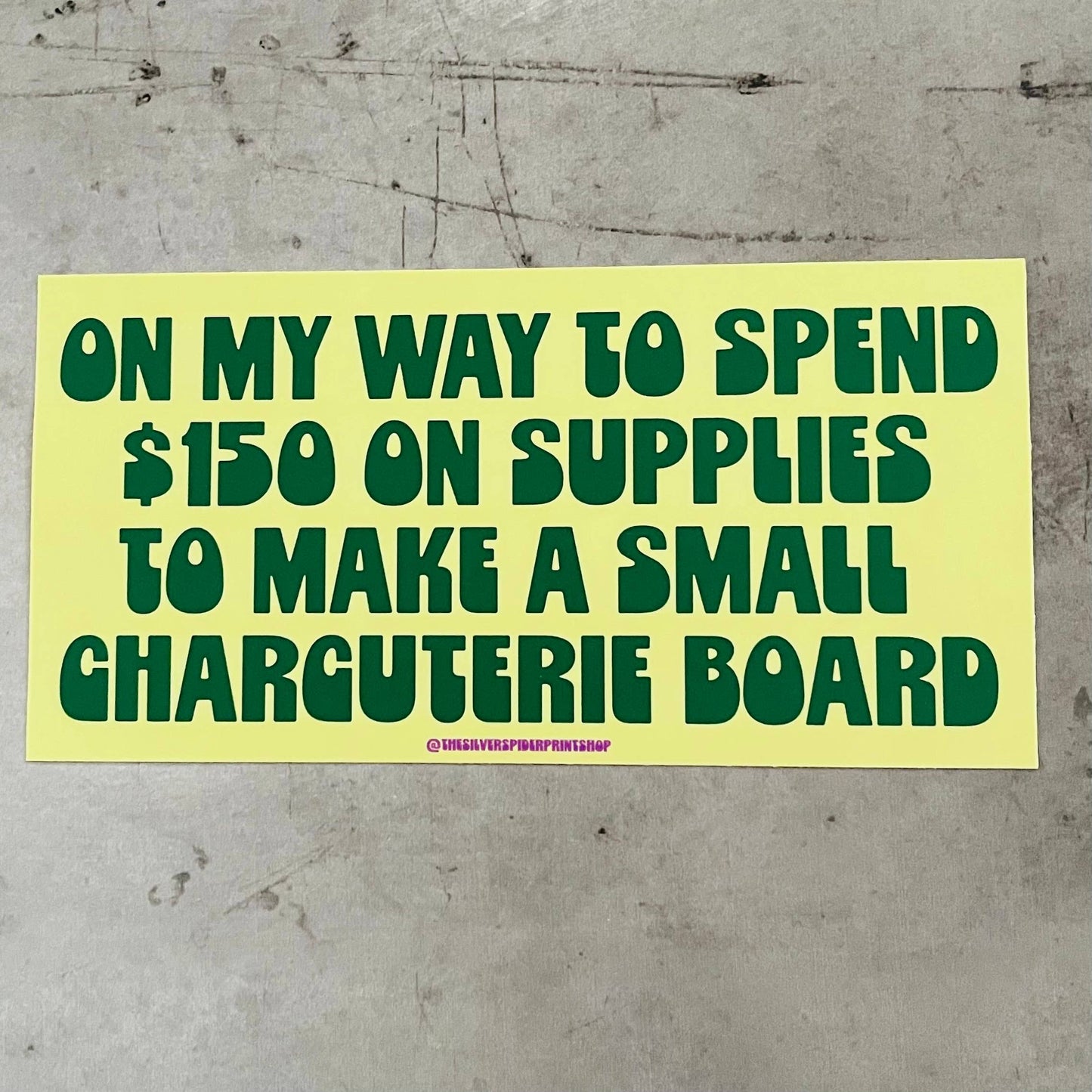 Yellow bumper stickerwith green text that reads "on my way to spend $150 on supplies to make a small charcuterie board" on a concrete surface