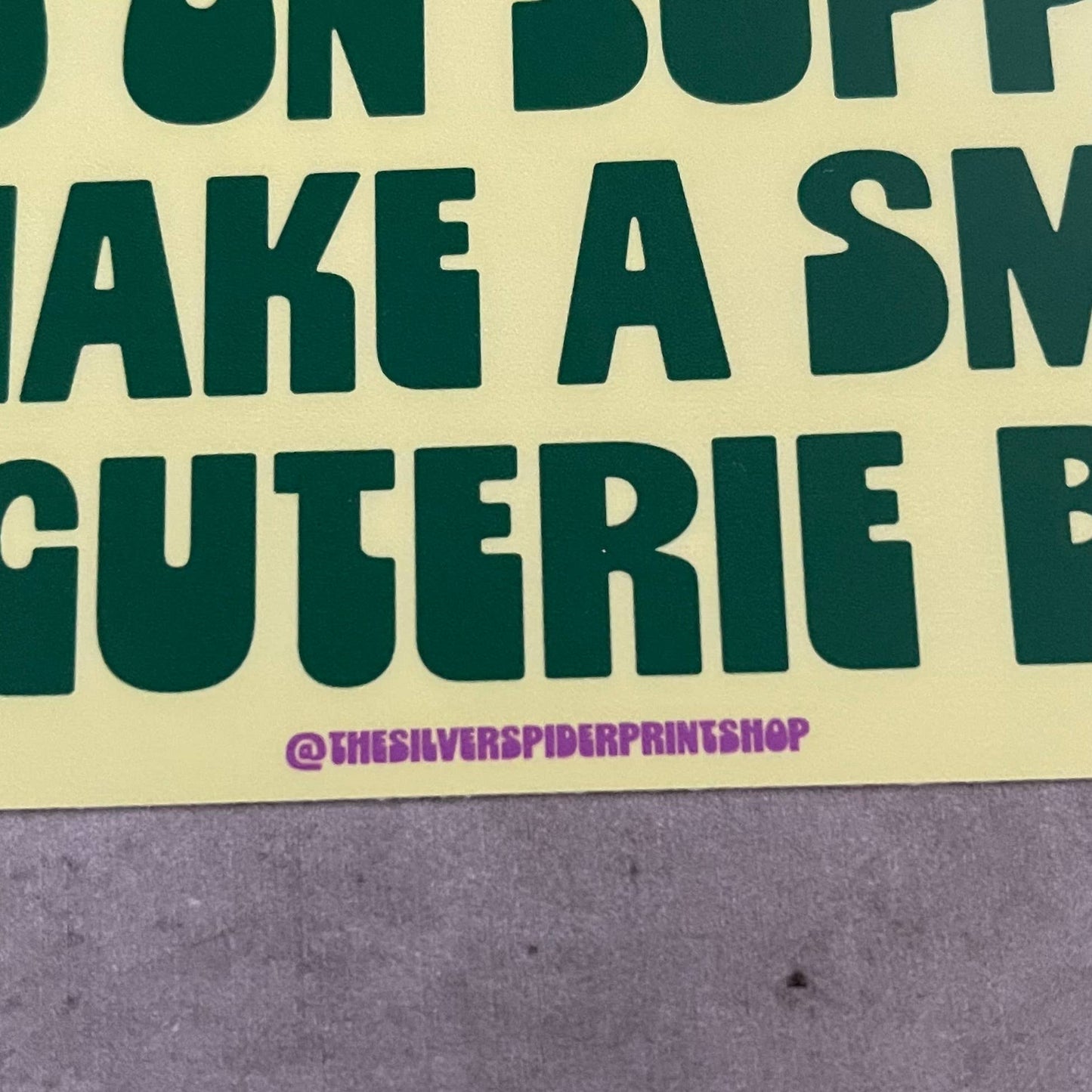 Yellow bumper stickerwith green text that reads "on my way to spend $150 on supplies to make a small charcuterie board" on a concrete surface