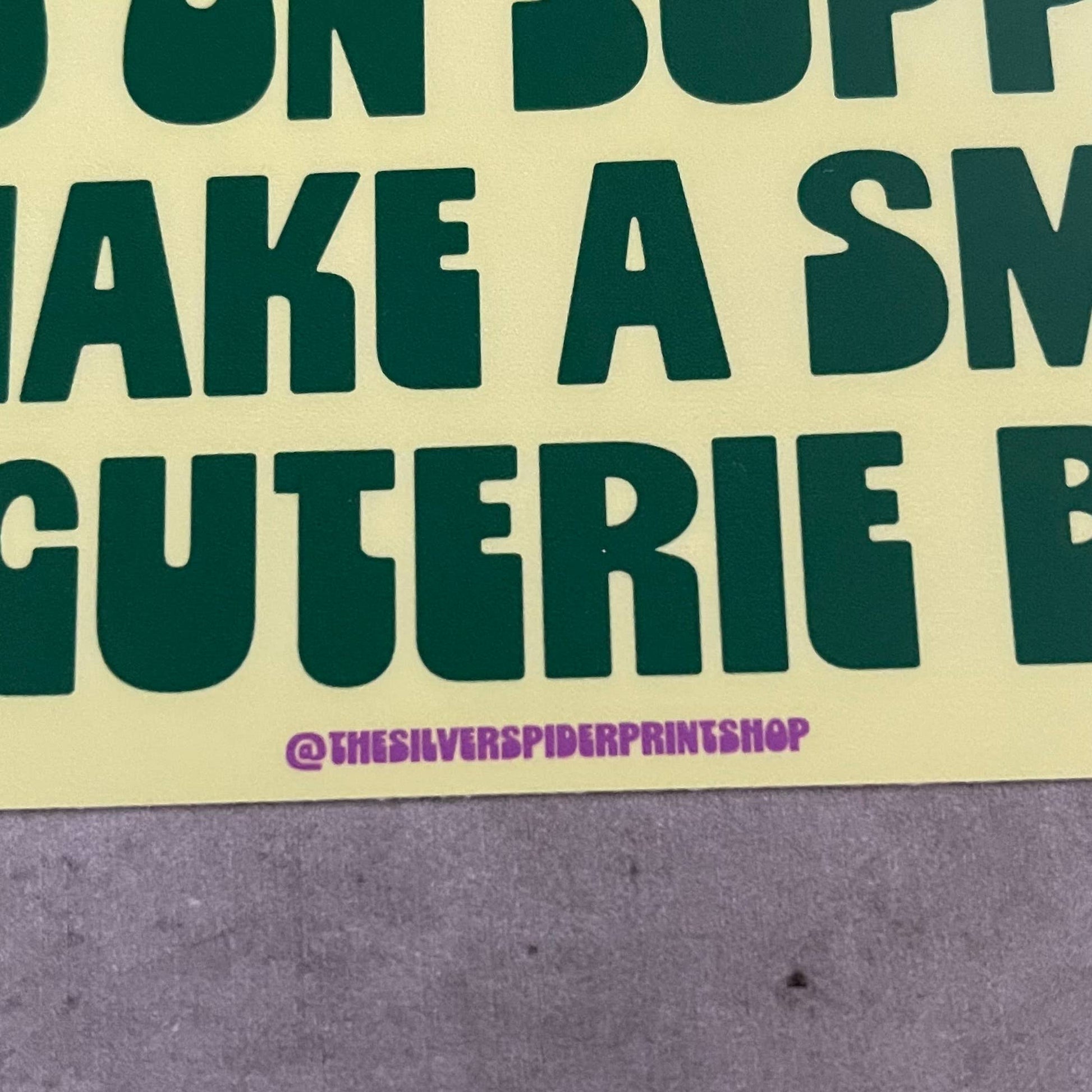 Yellow bumper stickerwith green text that reads "on my way to spend $150 on supplies to make a small charcuterie board" on a concrete surface