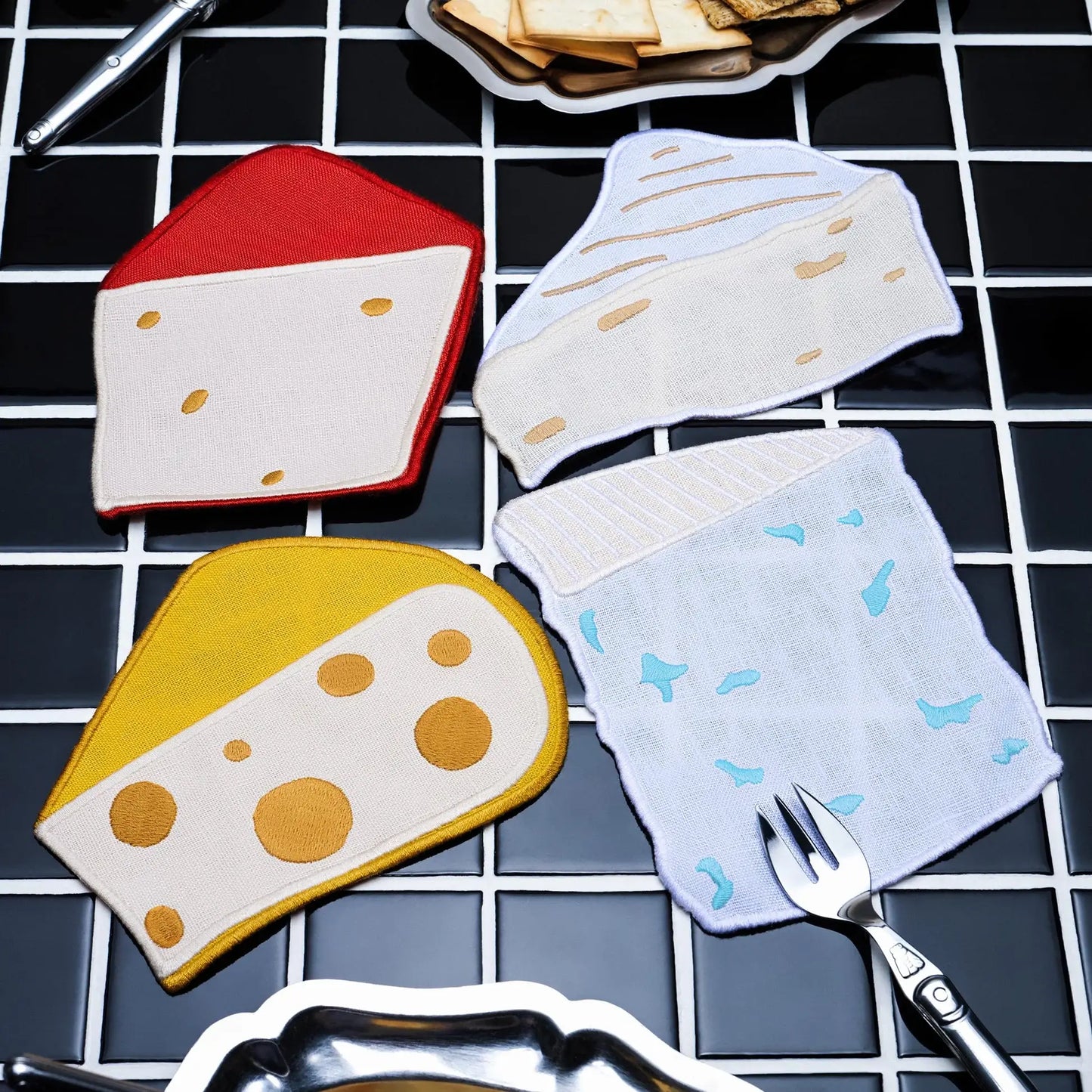 Cheese-shaped coasters/cocktail napkins on a checkered surface with a fork and crackers.