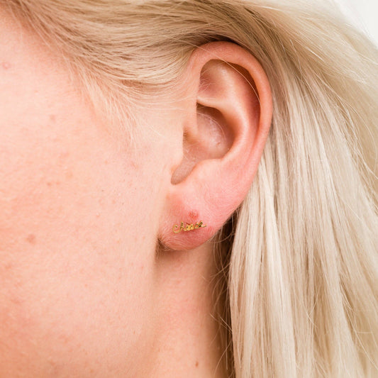 Single 14k gold plated stud earring -- reads "cheese" in handwritten script. Worn on ear for scale 