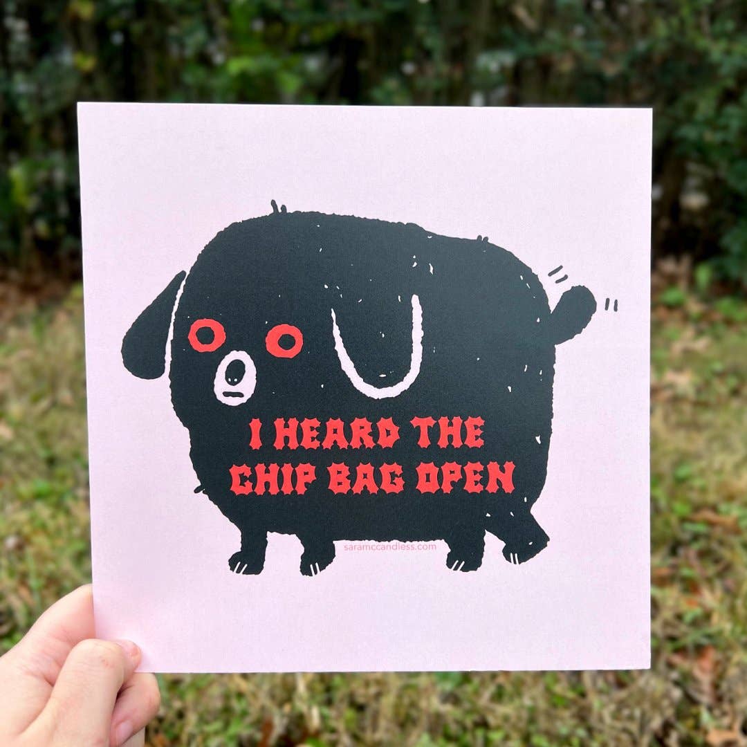 art print being held with image of a black dog with red eyes and red text that reads "I heard the chip bag open"