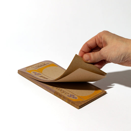 Hand peeling a piece of paper from the chocolate milk carton notepad pictured on a white background