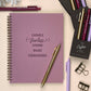 Purple notebook with motivational quote, pens, and flowers on a light background